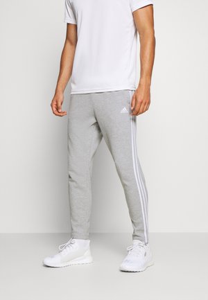Homme portant un pantalon de survêtement Adidas gris clair avec des bandes blanches sur les côtés, des baskets blanches et un t-shirt blanc à manches courtes, debout devant un fond uni.