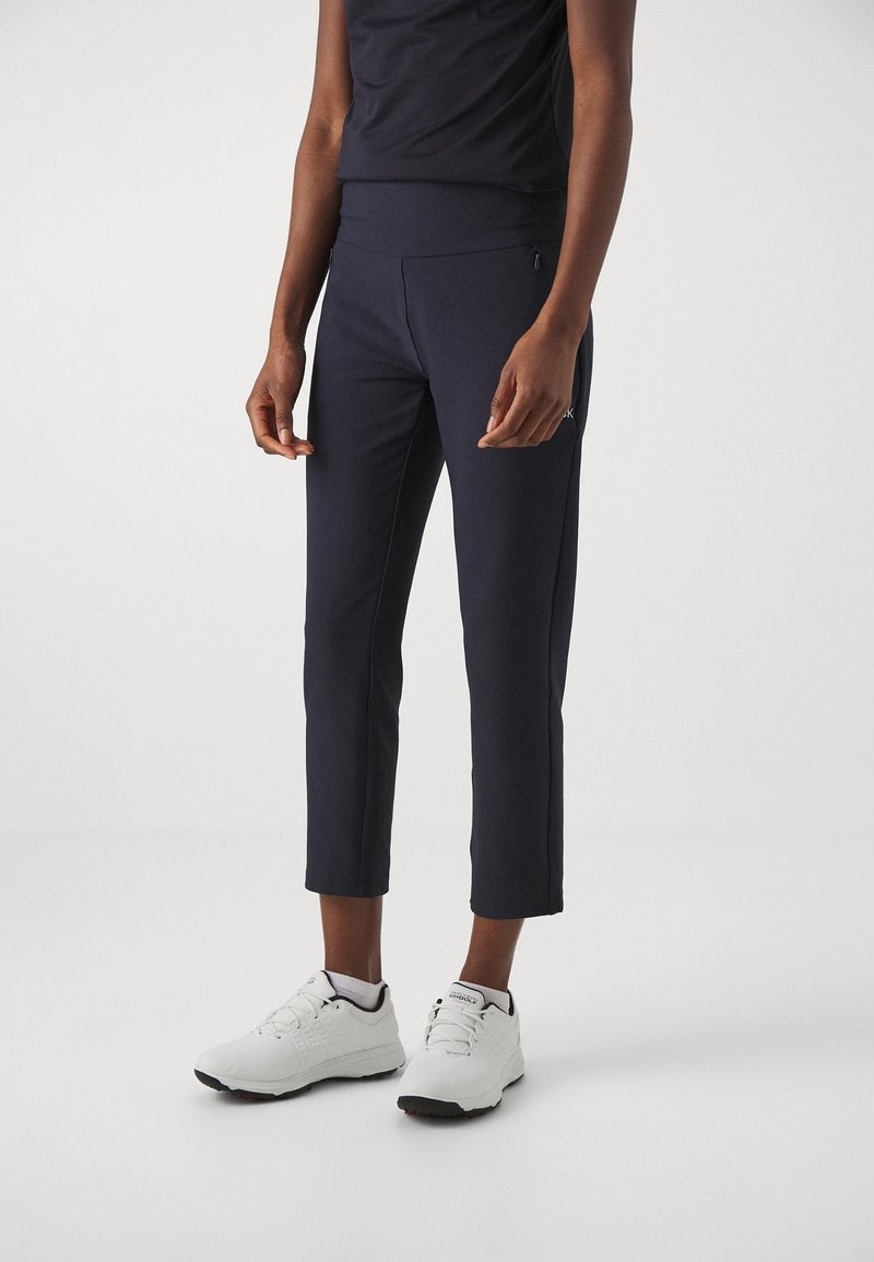 Personne portant un pantalon noir raccourci et des baskets blanches, debout devant un fond clair uni.