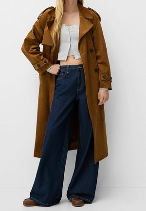 Kvinde iført brun ruskind trenchcoat, hvid kort knap-top, vidde ben mørkeblå jeans og brune sko, stående mod hvid baggrund.