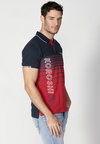 Polo tričko v námorníckej modrej a červenej farbe s horizontálnymi pruhmi. Má krátke rukávy, golier a výrazný nápis "KOROSHI" na prednej strane.