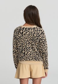 Donna con lunghi capelli castani che indossa un maglione leopardato e felpato e pantaloni corti beige, di spalle su uno sfondo chiaro uniforme.