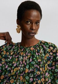 Bluse med blomsterprint i sort baggrund, med grønne, pink og orange blomster. Struktureret stof og detaljeret samling ved halsen.