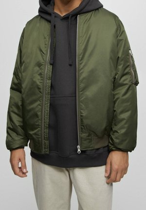 Homme portant un sweat à capuche gris foncé sous une veste bomber vert olive avec une poche zippée sur la manche, associé à un pantalon beige clair, debout devant un fond uni.