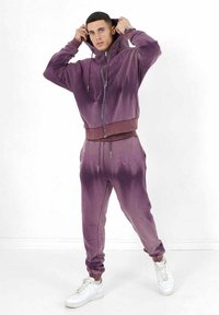 Lila Batik-Zip-Hoodie mit Kordelzugkapuze, kombiniert mit passenden Jogginghosen mit elastischem Bund und gestickten Akzenten. Weiße Sneakers.
