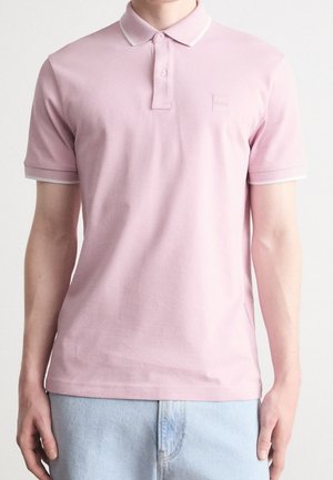 Mann trägt ein hellrosa kurzärmeliges Polohemd mit einem kleinen geprägten Logo auf der Brust und hellblaue Jeans.