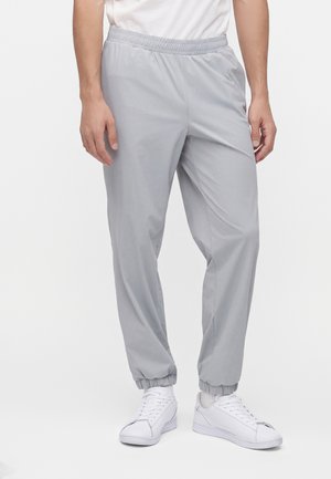 Homme portant un pantalon gris clair à taille et chevilles élastiques, des baskets blanches et un haut blanc, debout sur un fond blanc uni.