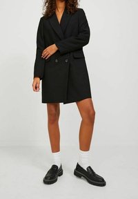 Svart dubbelknäppt kavajklänning med långa ärmar, slagkrage och framfickor. Matchas med svarta loafers och vita strumpor.