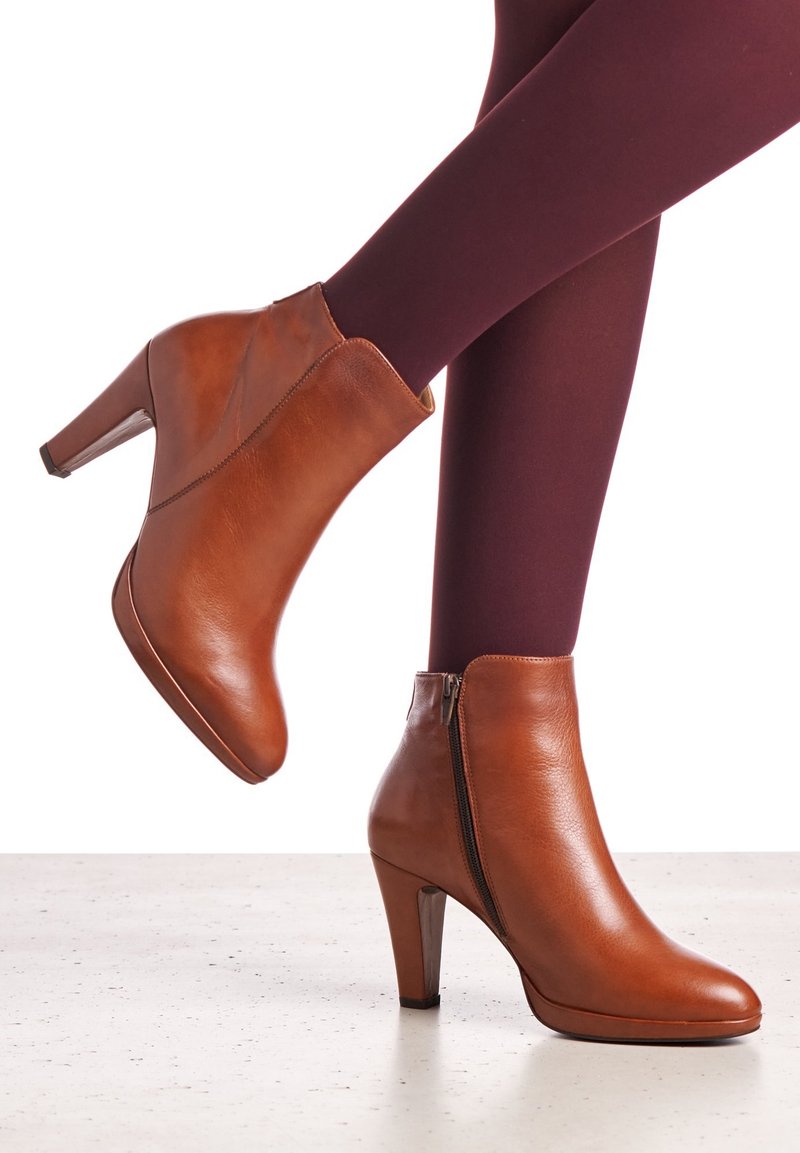 Femme portant des bottines en cuir marron avec des fermetures éclair, associées à des collants violet foncé, un pied levé derrière l'autre.