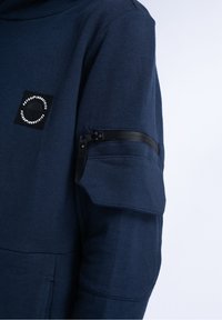 Le sweat-shirt bleu marine présente une poche zippée sur la manche, des coutures renforcées et un patch logo circulaire sur le haut du bras.