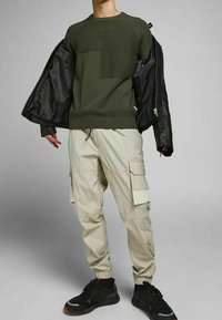 Pull texturé vert olive avec un motif côtelé, associé à un pantalon cargo beige clair avec plusieurs poches et des poignets élastiques. Baskets noires.