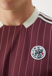 Maillot de sport rayé bordeaux avec un col ras du cou. Il présente un patch brodé circulaire noir et blanc représentant une silhouette avec un accent rouge.