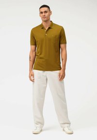 Korte mouwen, mosterdkleurige poloshirt met een kraag, zak en contrasterende blauwe afwerking. Gepaard met lichtgekleurde, losse broek en sneakers.