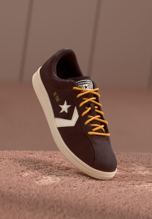 Mørkebrun semsket sneakers med hvit stjerne-logo og detaljer, med oransje snører og kremfarget såle. Teksturert overflate, lavt design.