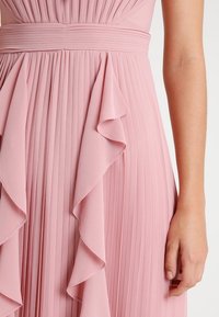 Roze geplooid jurkje met ruches op de taille. Glad stof met verticale textuur, toont een close-up van het kledingstuk en een hand.