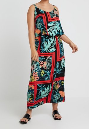 Kvinde iført en ærmeløs, ankel-lang kjole med farverige tropiske blade og blomster mønstre indrammet af røde geometriske linjer, kombineret med sorte slip-in sandaler.