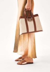 Osoba u prozirnoj bež haljini, drži torbu s ručkama od smeđe kože i bež platna, nosi usklađene smeđe i bež natikače.