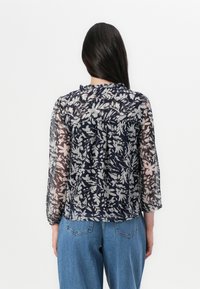 Una donna con capelli lunghi indossa una blusa trasparente con un motivo floreale, mettendo in mostra il design del retro. Abbina la blusa a un paio di jeans blu.