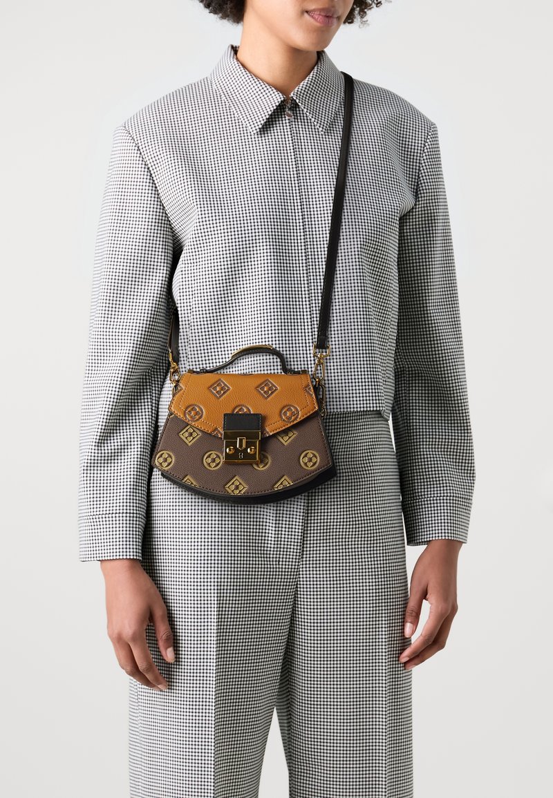 Femme portant un petit sac bandoulière marron et beige à motif avec une fermeture dorée sur une tenue à carreaux noire et blanche.