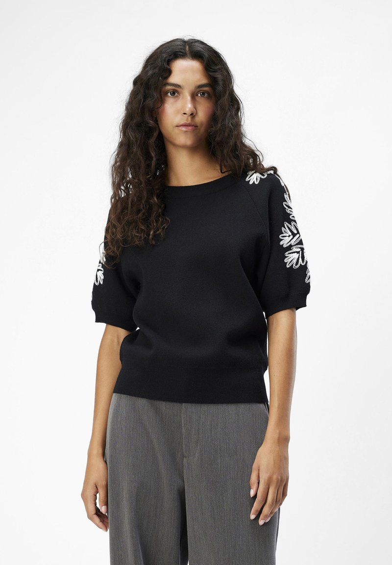 Jersey negro con mangas cortas abullonadas, que presenta bordados florales blancos en los hombros. Textura suave, entallado en la cintura.