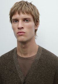Brown textured cardigan over a brown shirt, featuring a V-neck and button details, worn by a model with short blond hair against a light background.