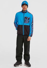 Junger Mann, der eine blau-schwarze Zip-Jacke mit roten Akzenten, schwarze Hosen, schwarze Stiefel und eine blaue Mütze vor einem weißen Hintergrund trägt.