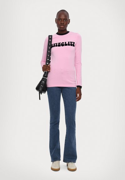 Pink long-sleeve shirt with black ribbed collar and bold text, paired with flared blue denim jeans. Accessory is a black shoulder bag.