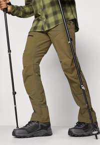Pantalons de randonnée kaki avec genoux articulés et poches zippées, associés à des chaussures de randonnée noires robustes, complétés par des bâtons de trekking.