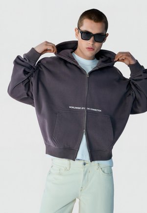 Ung mand iført sorte solbriller løfter hætten på en mørk lynlåshættetrøje med hvid tekst, kombineret med lyse bukser.