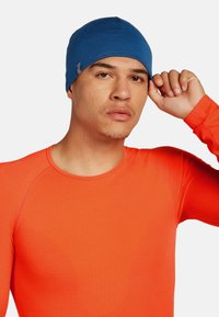 Man wearing a fitted blue beanie and bright orange long-sleeve shirt, adjusting the beanie with his right hand against a plain background.