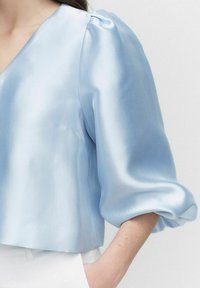 Ljusblå satinstopp med djup V-ringning, puffärmar och slät textur. Kort längd med en ren avslutning vid fållen.