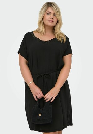 Kvinde iført en sort knælængde kjole med blonder ved halsudskæringen og bånd i taljen, holder en lille sort håndtaske foran en ensfarvet baggrund.