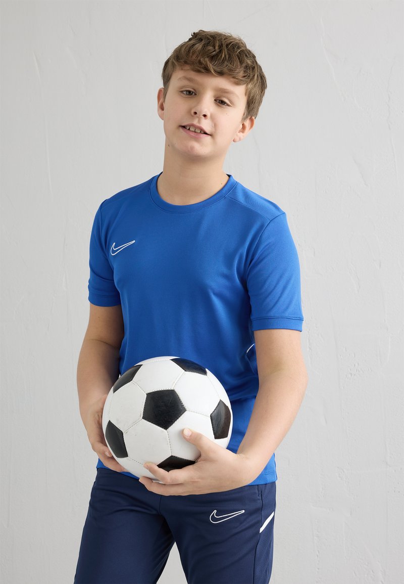 Camiseta deportiva azul de manga corta con el logo de Nike, combinada con pantalones cortos azul marino, sosteniendo un balón de fútbol blanco y negro frente a un fondo claro.