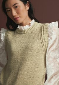 Strikket sweatervest i lys oliven grøn med ribbet halsudskæring, parret med en hvid bluse med flæser, der har blomster mønstre på ærmerne.