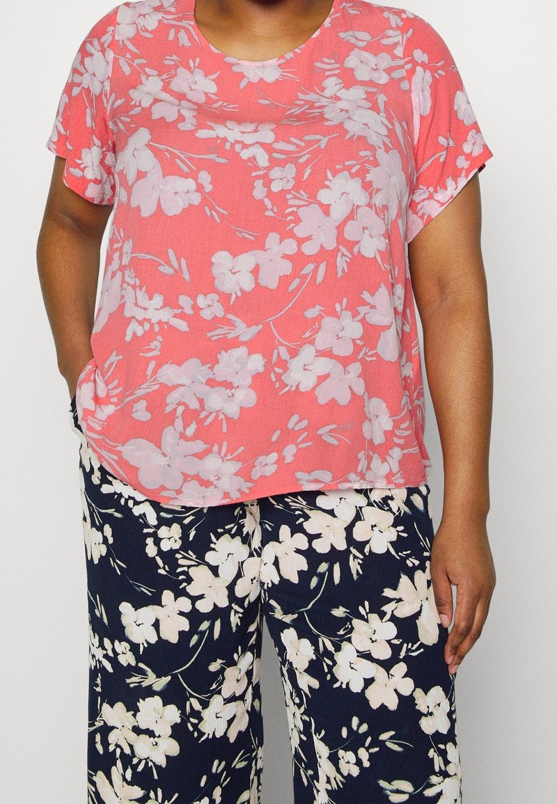 Blouse à manches courtes corail avec un motif floral pâle, associée à un pantalon large marine orné de grandes fleurs blanc cassé. Texture de tissu douce.