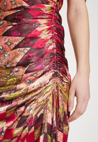 Tissu froncé à motifs multicolores rouge vif sur une robe avec des motifs floraux et cachemire, à côté d’un bras et d’une main pâles.