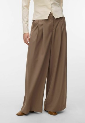 Frau trägt hoch sitzende, weite braune Hose, kombiniert mit beigem, geknöpftem Oberteil und braunen Schuhen, steht vor einem schlichten Hintergrund.