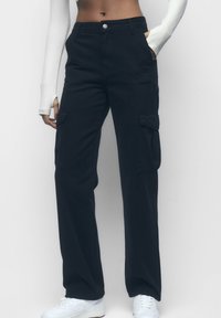 Personne portant un pantalon cargo noir taille haute avec des poches latérales, un haut à manches longues blanc et des baskets blanches, se tenant contre un fond uni.