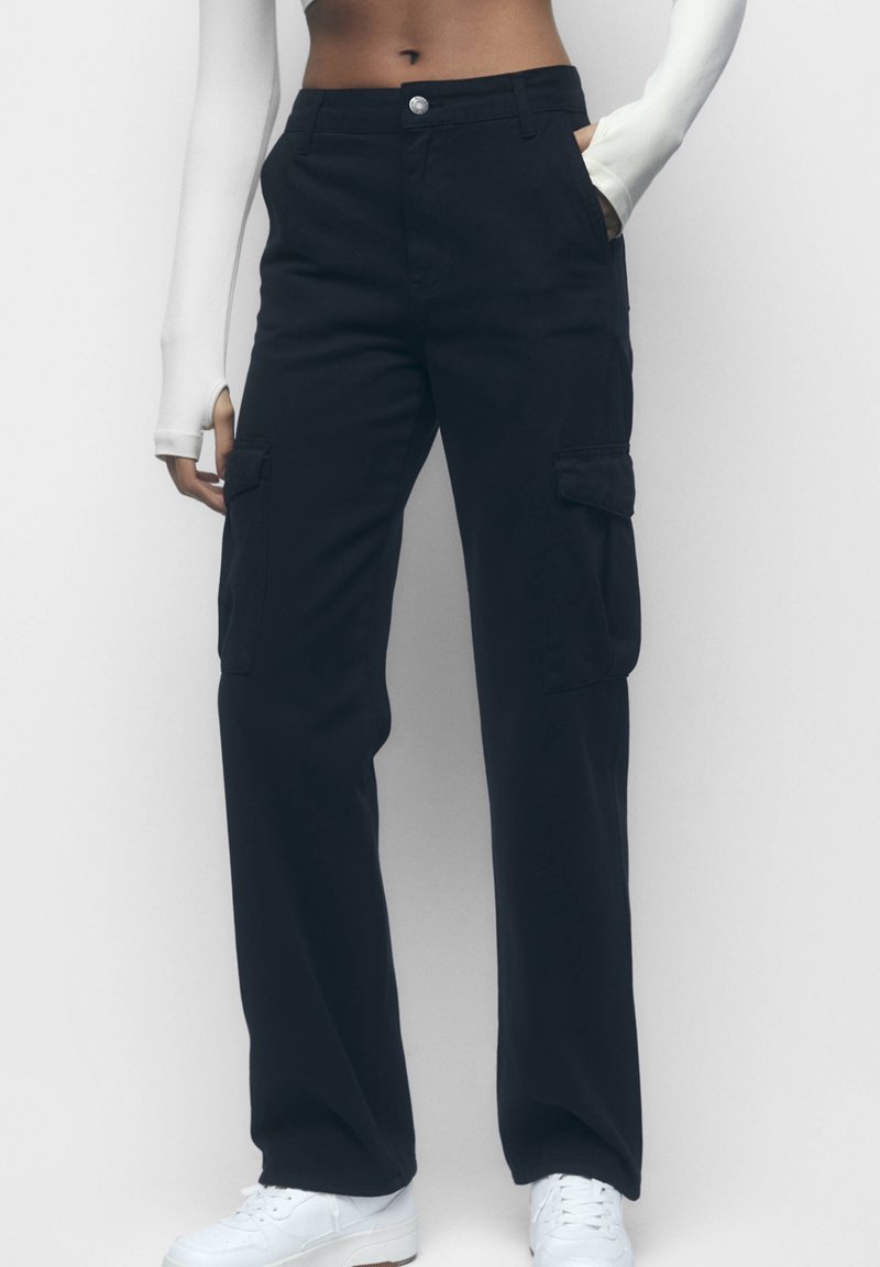 Personne portant un pantalon cargo noir taille haute avec des poches latérales, un haut à manches longues blanc et des baskets blanches, se tenant contre un fond uni.