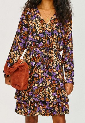 Kobieta w sukience z długim rękawem w kwiaty w kolorach fioletu, pomarańczu i kremowym, trzymająca po bokach zamszową torebkę w kolorze rdzy.