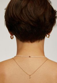 Pendientes de oro y dos collares en capas. Uno de los collares cuenta con perlas, mientras que el otro tiene una cadena de oro con un colgante en forma de "P". Cabello corto y oscuro.