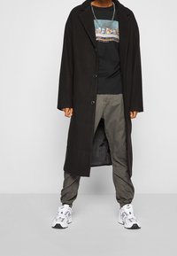 Lange zwarte jas met een kentkraag, knoopsluiting, gedragen over een graphic t-shirt, gecombineerd met olijfgroene joggers en witte sneakers.