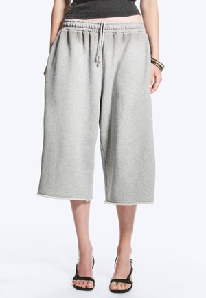 Femme portant un pantalon de survêtement ample gris clair mi-genou avec taille à cordon et sandales noires à talons ouverts, debout sur fond blanc.