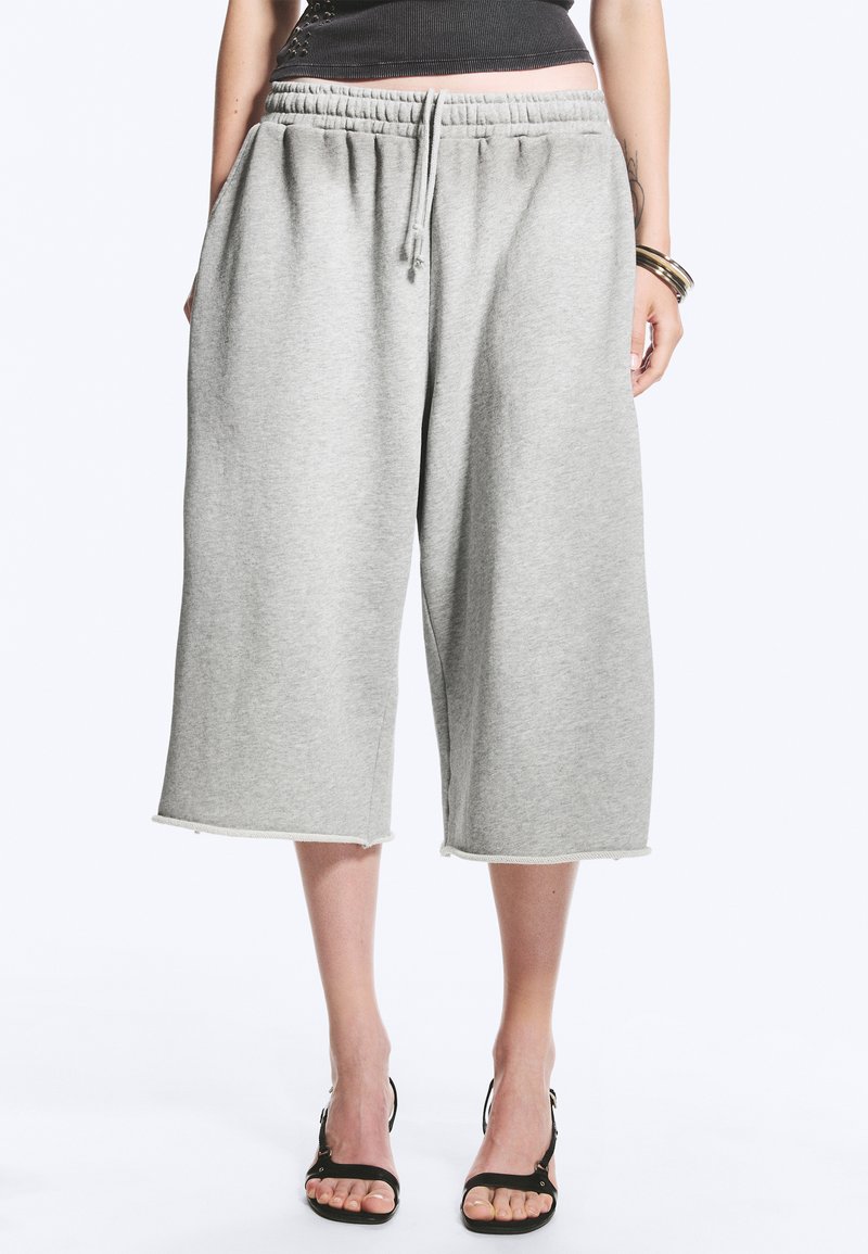 Femme portant un pantalon de survêtement ample gris clair mi-genou avec taille à cordon et sandales noires à talons ouverts, debout sur fond blanc.