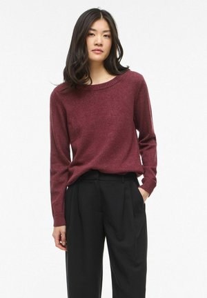 Ung kvinde med langt mørkt hår iført en mørkerød langærmet sweater og sorte bukser med høj talje, stående med den ene hånd i lommen.