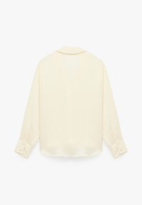 Krämfärgad blus med långa ärmar, subtil textur och en ryggsömm. Knappar vid ärmsluten och en rundad nederkant fulländar designen.