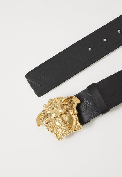Cinturón de cuero negro con una superficie texturizada y una prominente hebilla en tono dorado con la cabeza de Medusa, que presenta detalles de marca grabados.