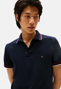 Polo azul marino con cuello y detalles en las mangas a rayas en rojo y blanco. Cuenta con dos botones en el escote y un pequeño logotipo en el pecho.