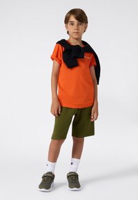 Camiseta naranja, sudadera negra caída sobre los hombros, pantalones cortos verdes oliva, calcetines deportivos blancos y zapatillas verdes con acentos.