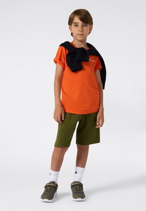 Orangenes T-Shirt, schwarzer Hoodie über die Schultern gelegt, olivgrüne Shorts, weiße Sportsocken und grüne Sneakers mit Akzenten.