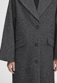 Cappotto houndstooth in bianco e nero, design oversize, ampie risvolti, chiusura frontale con bottoni, due tasche laterali, tessuto testurizzato.